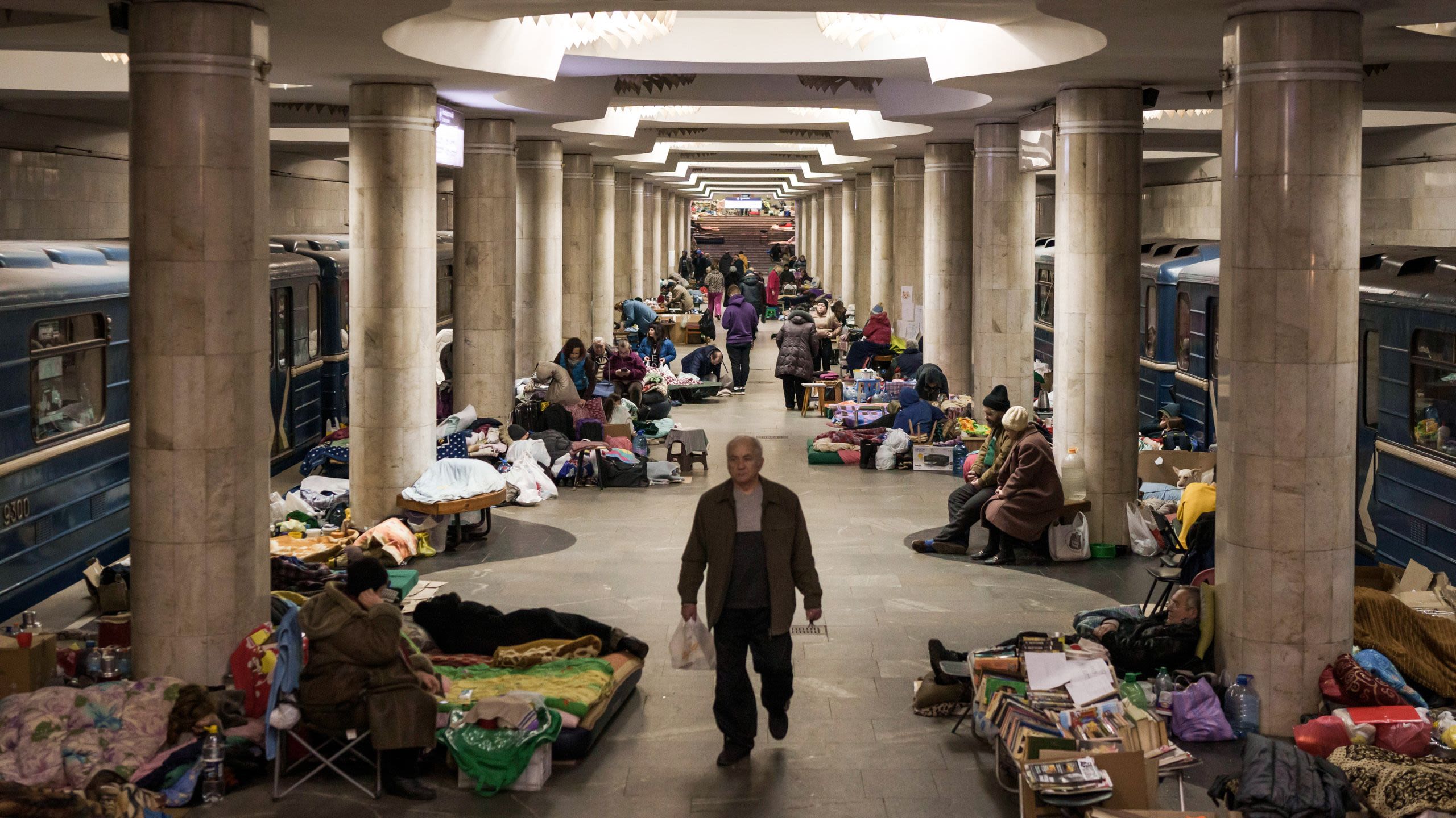 People mill about an underground subway platform that is adorned with blankets, boxes, and makeshift beds.