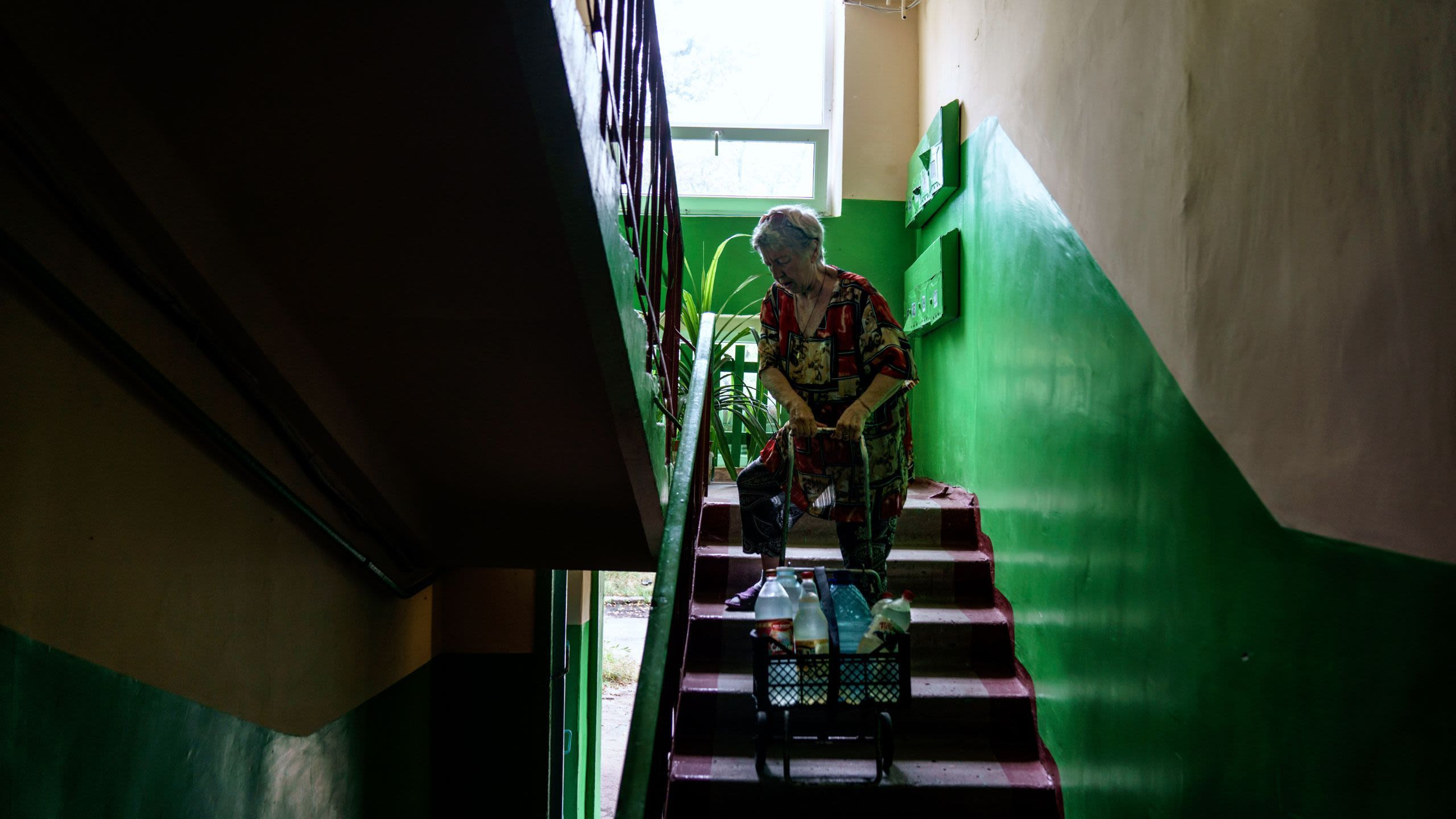 An elderly woman ascends a staircase pulling a shopping cart full of bottles behind her.