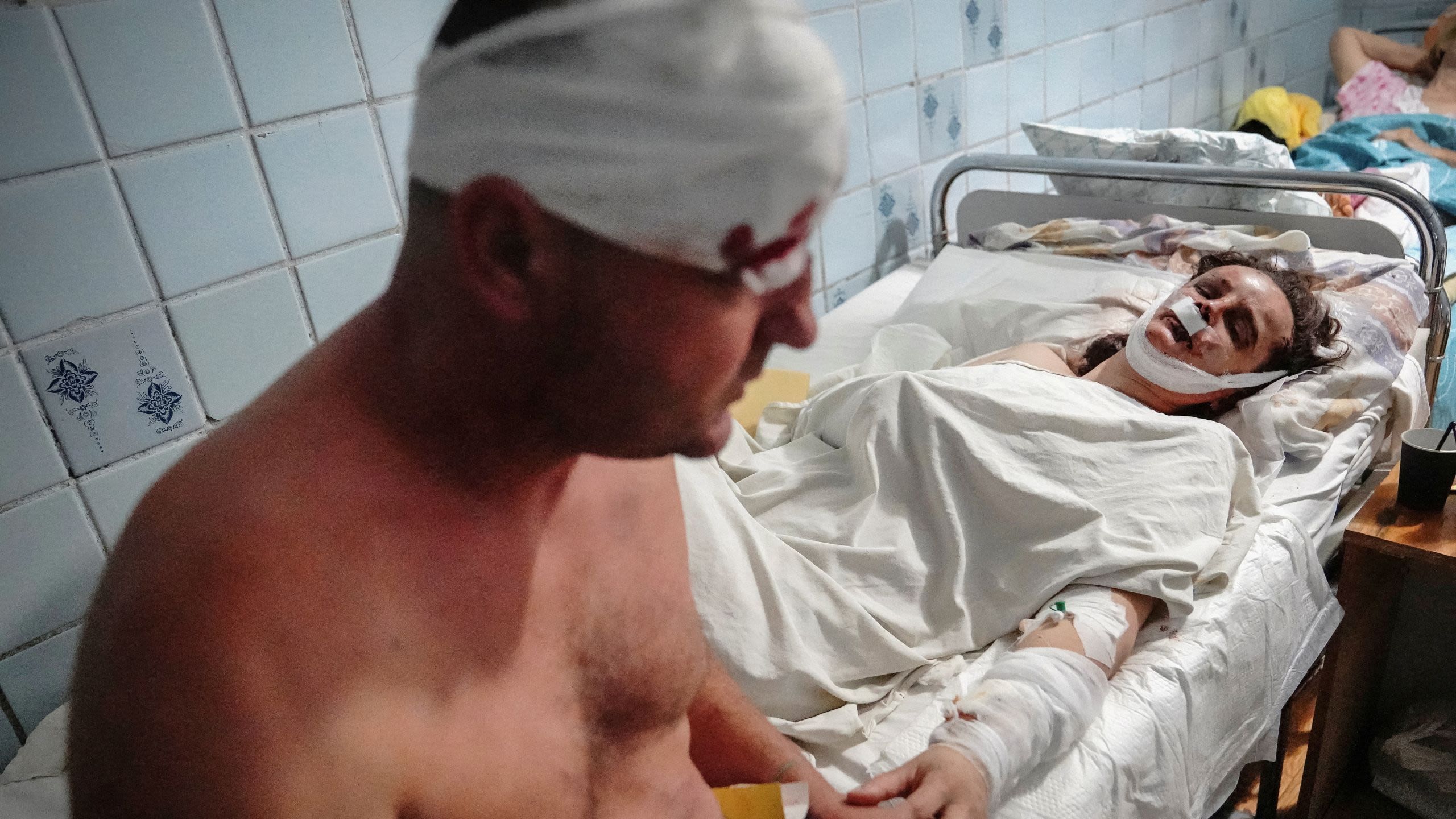 A shirtless man with a bloodied bandage wrapped around his head holds the hand of bandaged woman lying in a gurney in a hospital hallway.