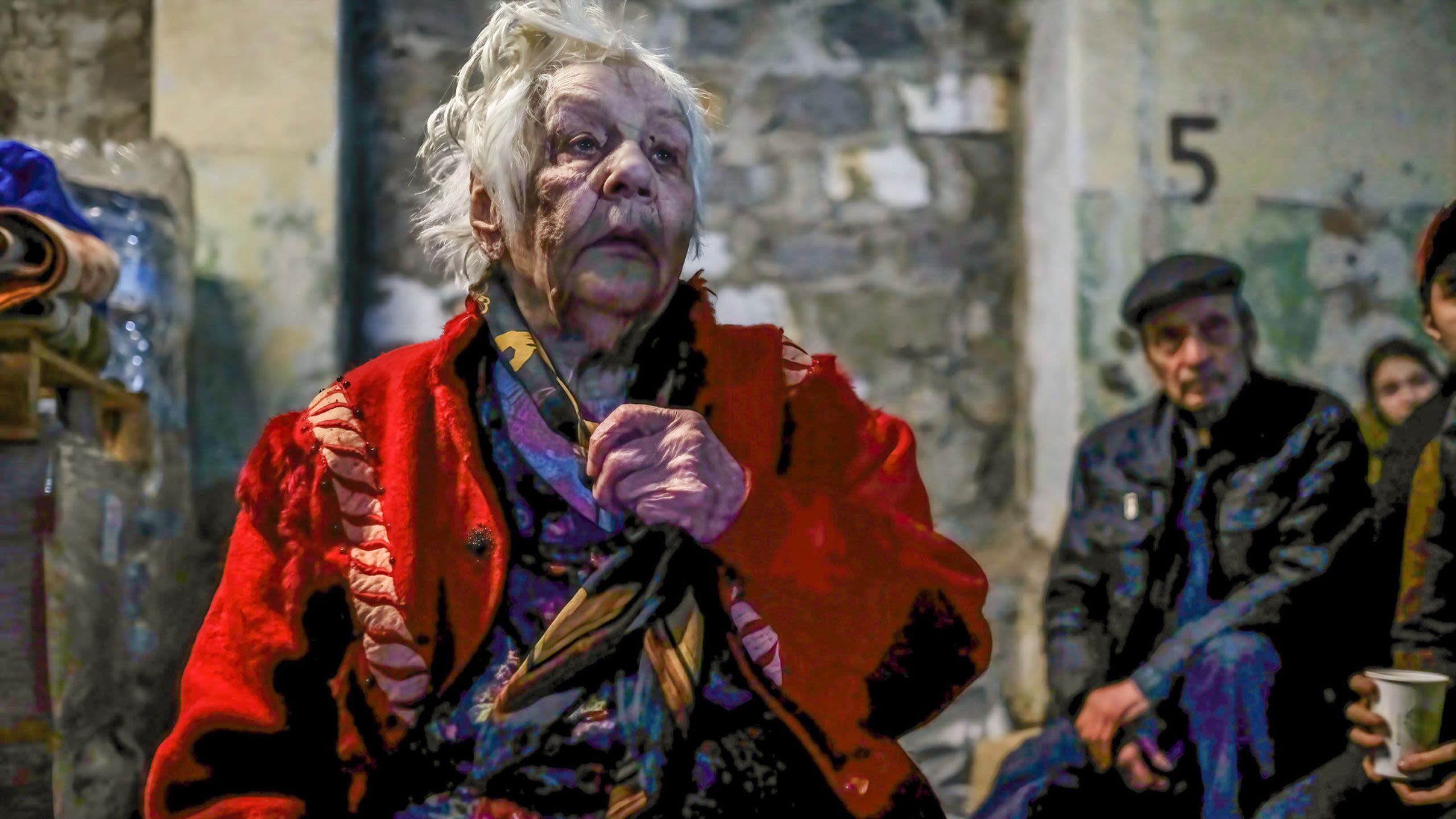 An unkempt and visibly exhausted elderly woman clutches the scarf around her neck and stares off into the distance.