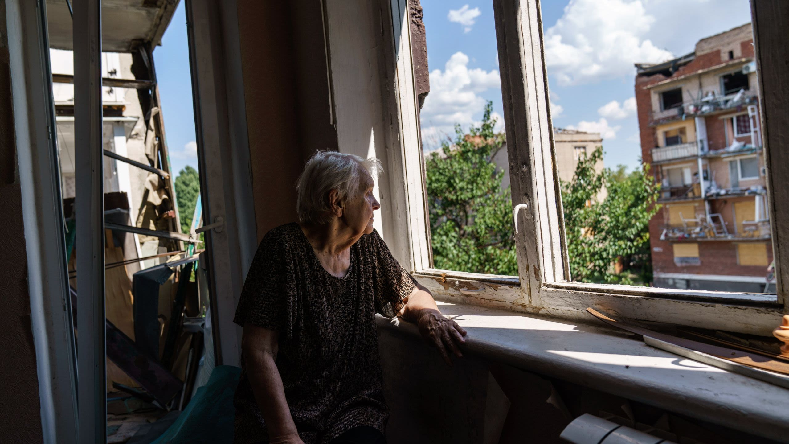 An elderly woman gazes out the window from her damaged apartment.