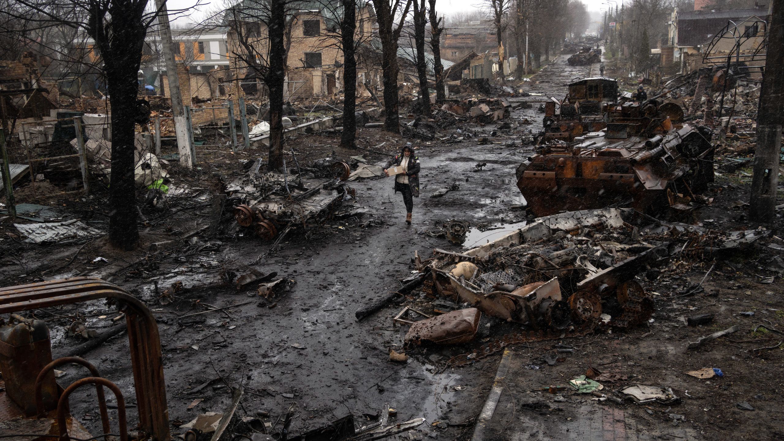 A woman carrying a box walks past a burned Russian tank.