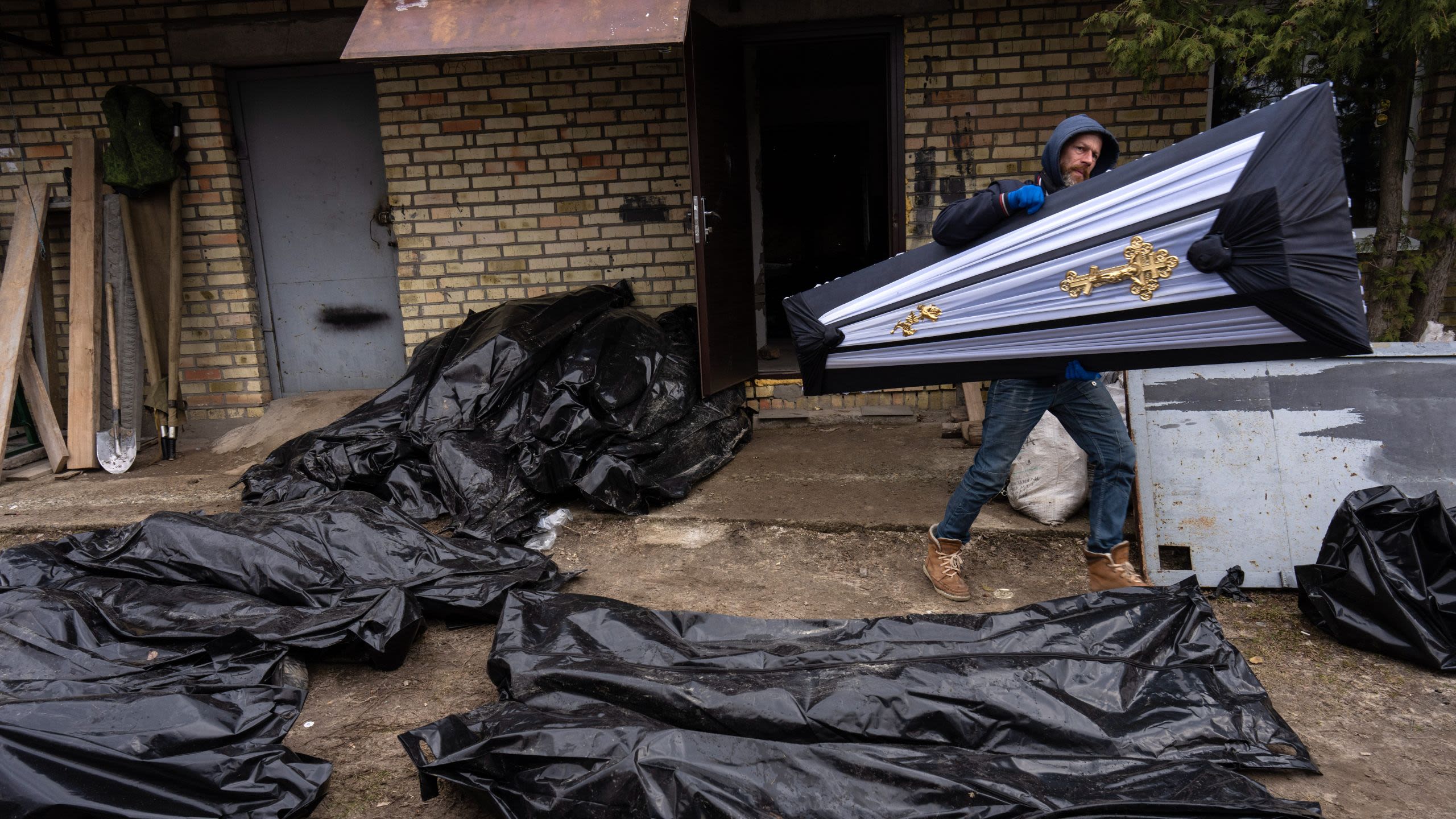 A man carries a coffin past a group of body bags on the ground.