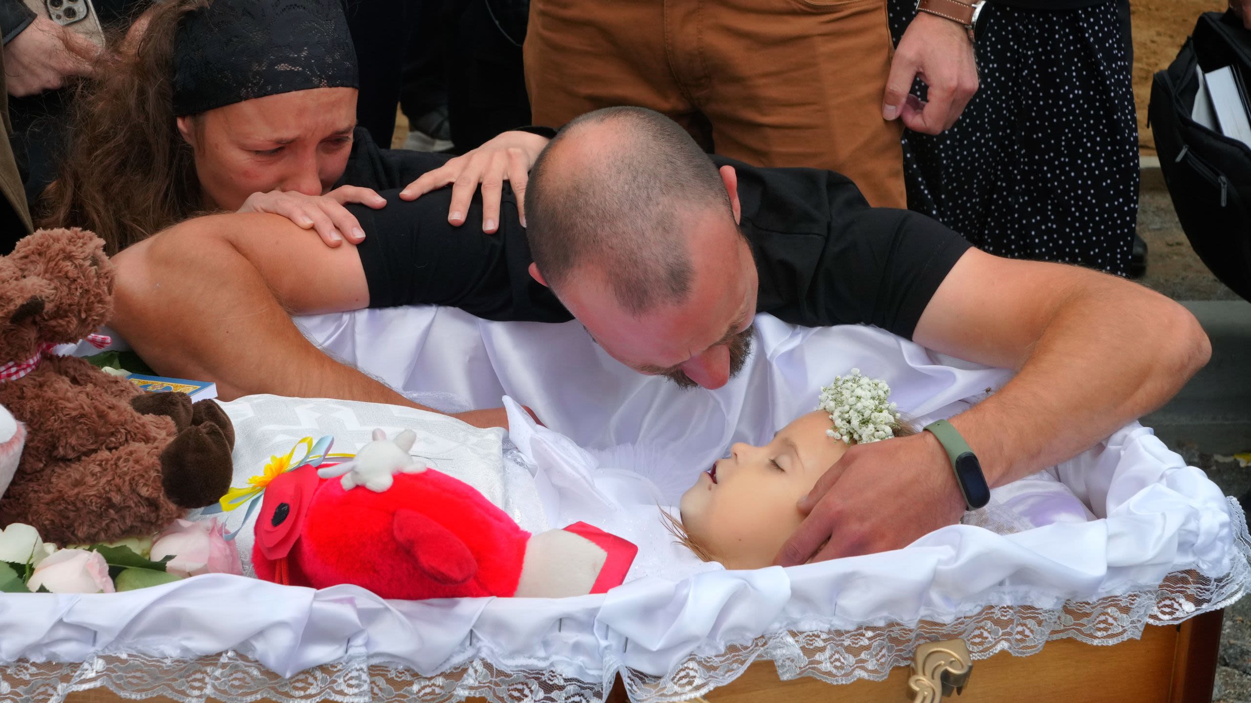 A sobbing man leans over an open casket, touching the cheek of the small girl inside it.