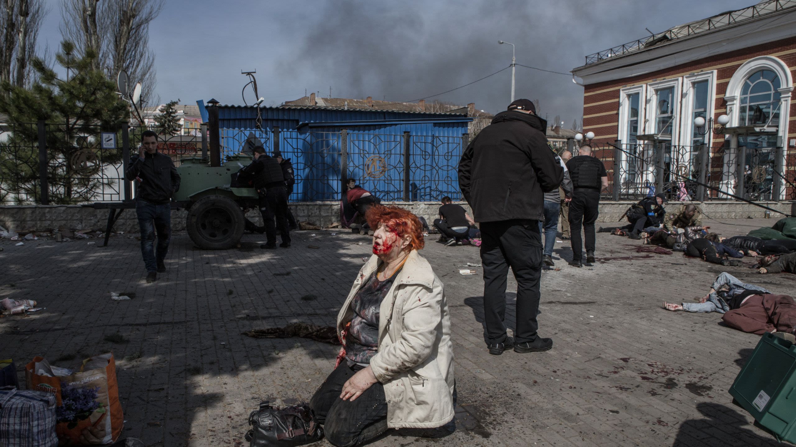 A bloodied woman kneels on the ground surrounded by bodies as black smoke fills the sky behind her.