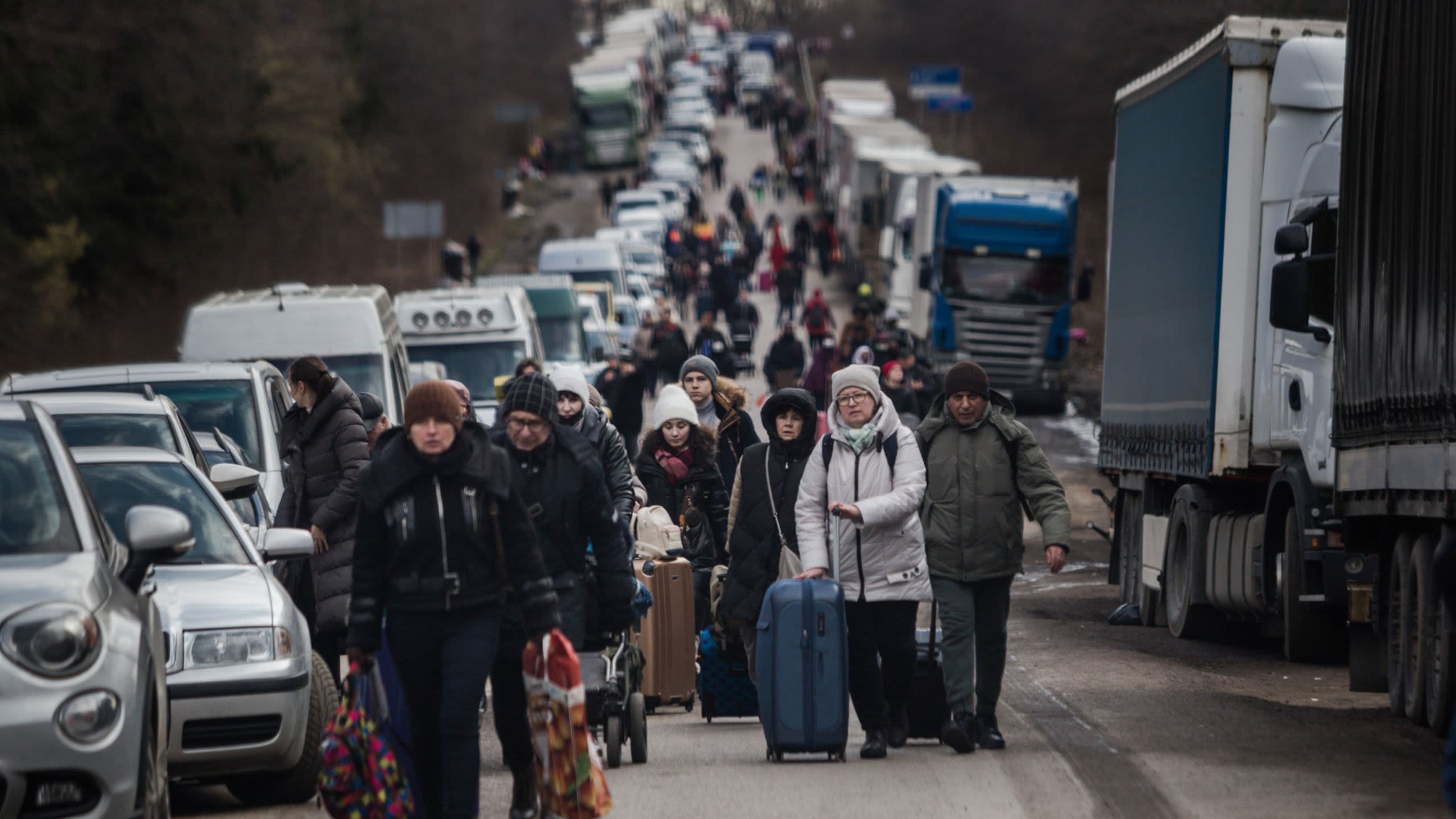A long line of people dressed in winter coats walk down a road carrying suitcases and other bags.