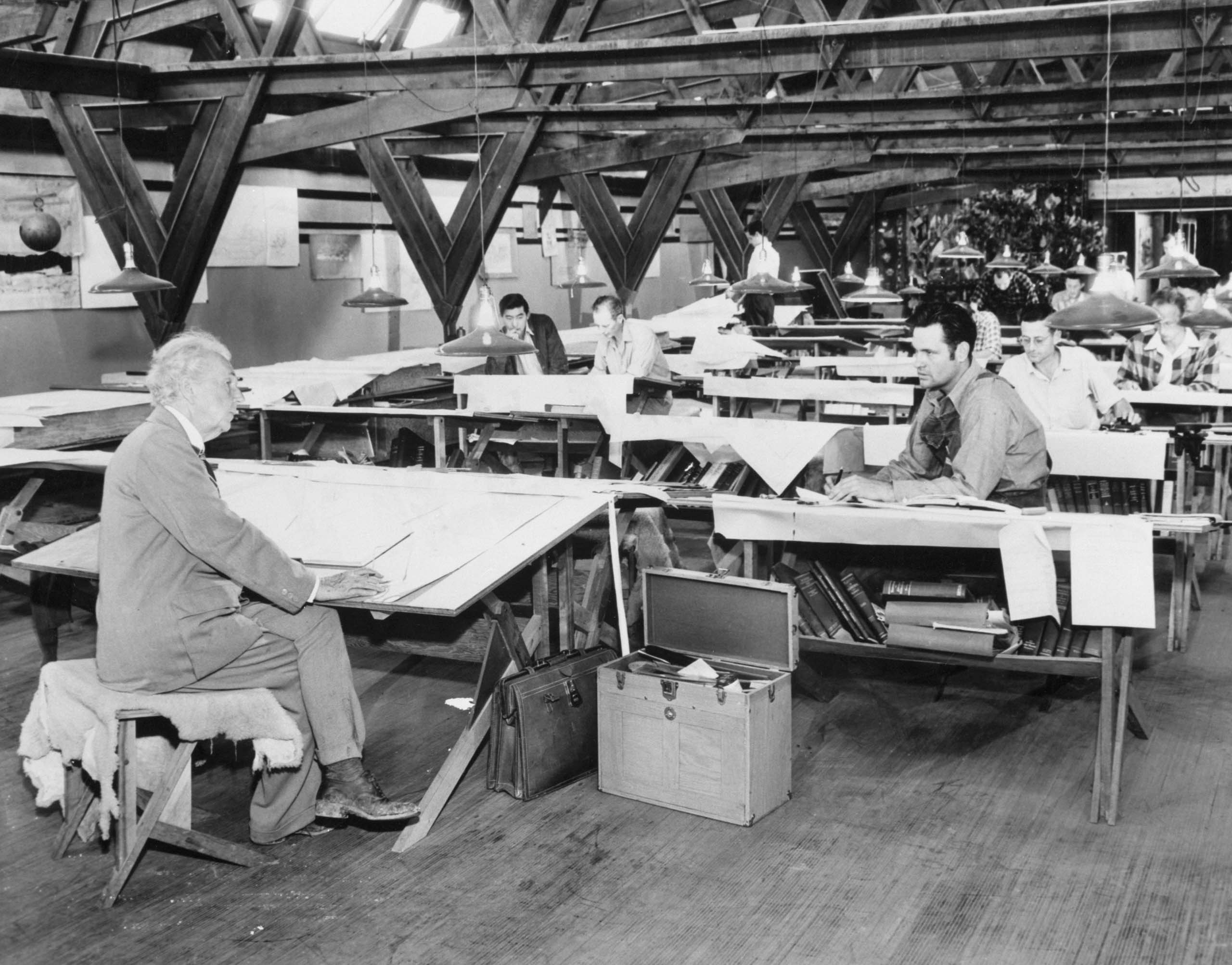 A photo shows Frank Lloyd Wright seated at a drafting table in the foreground, with several other men working at tables behind him in a large, open studio (© Bettmann/Getty Images)