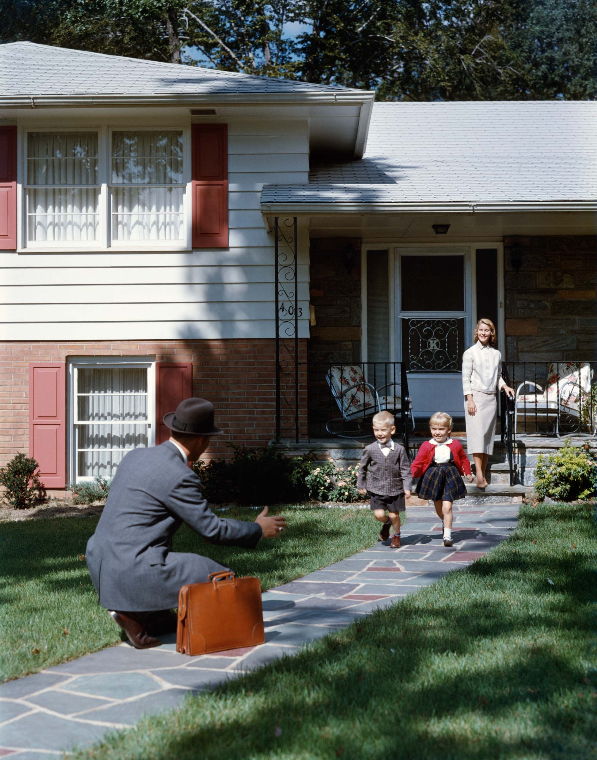 A man in a suit kneels on a stone walkway to greet a young boy and girl running toward him, with a woman standing on the porch of a split-level home in the background (© H. Armstrong Roberts/ClassicStock/Getty Images)