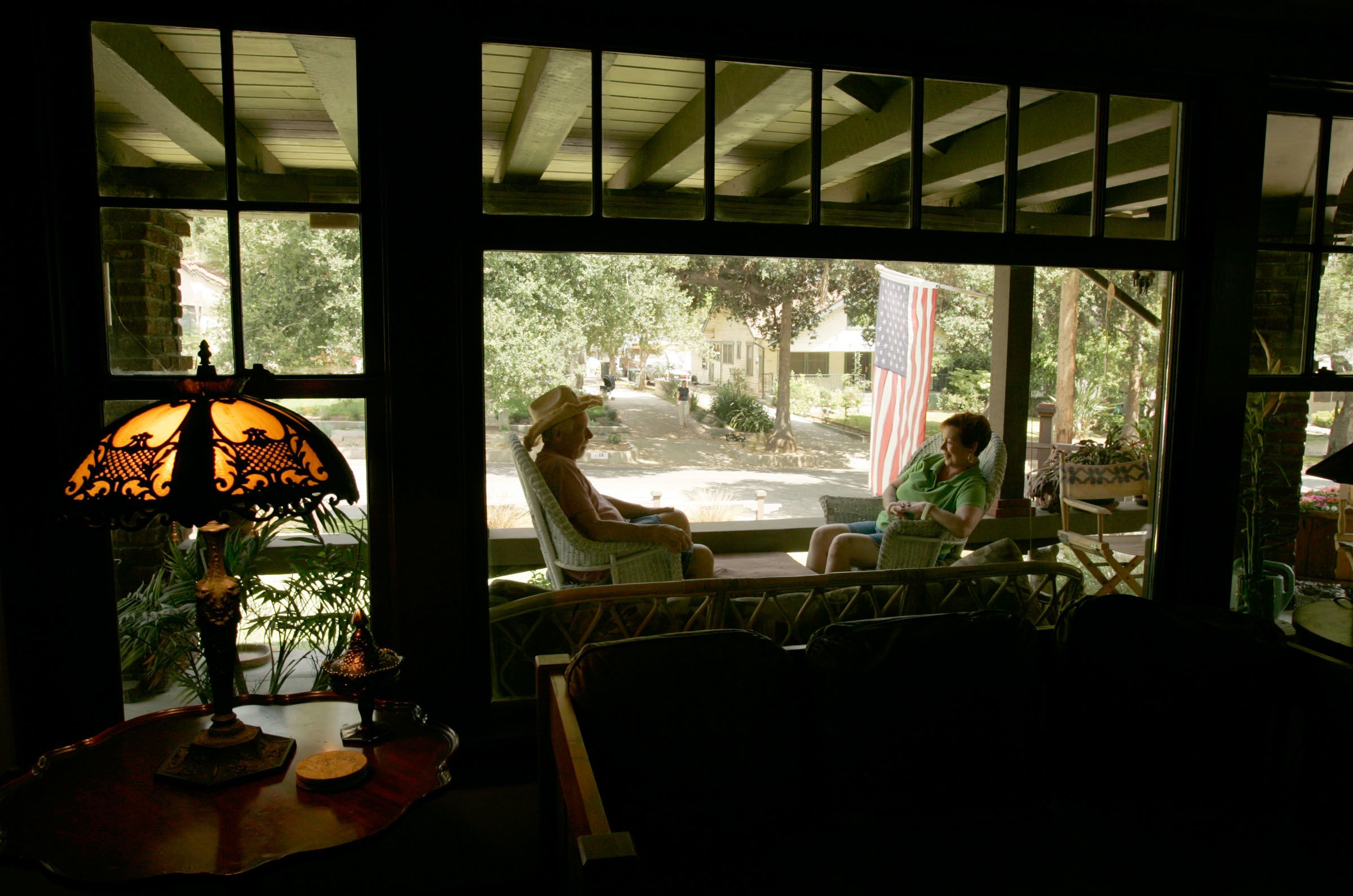A view through a window shows two people sitting in chairs on a porch, facing each other, while an American flag hangs in the background (© Mark Boster/Los Angeles Times/Getty Images)