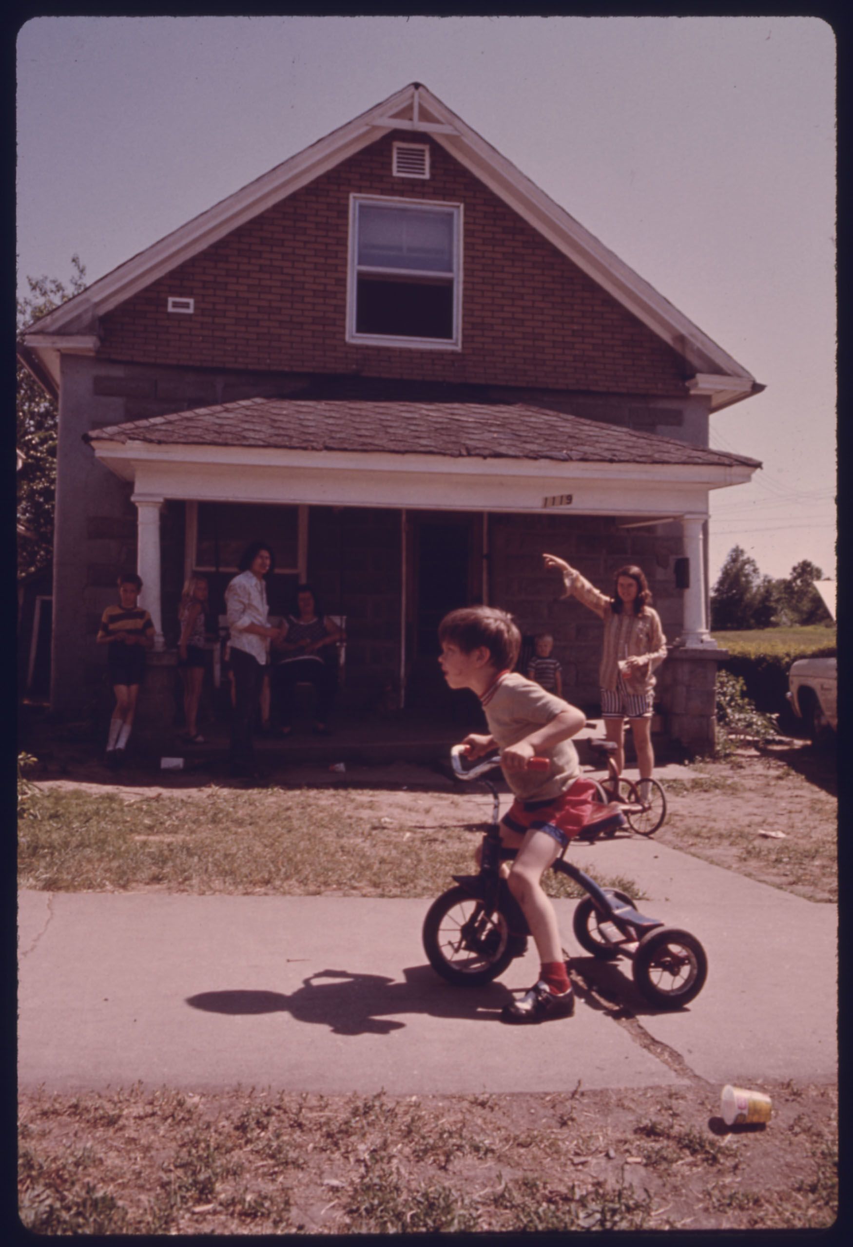 A young boy rides a tricycle on a sidewalk in front of a two-story house while several people stand on the front porch and in the yard behind him (National Archives/Environmental Protection Agency/Kenneth Paik)