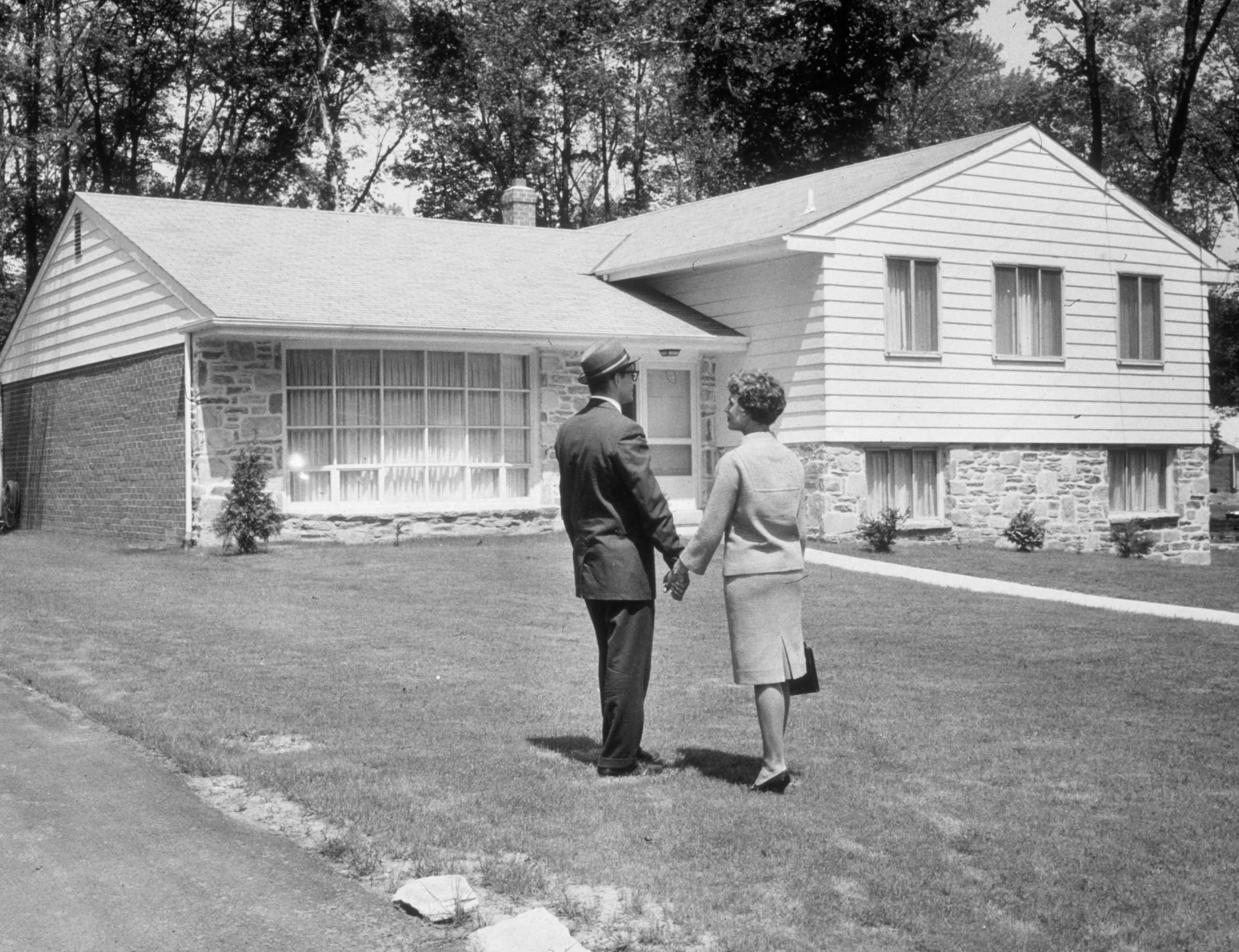 A photo shows a man and a woman standing hand-in-hand in a grassy yard, facing a split-level ranch-style house (© Harold M. Lambert/Getty Images)