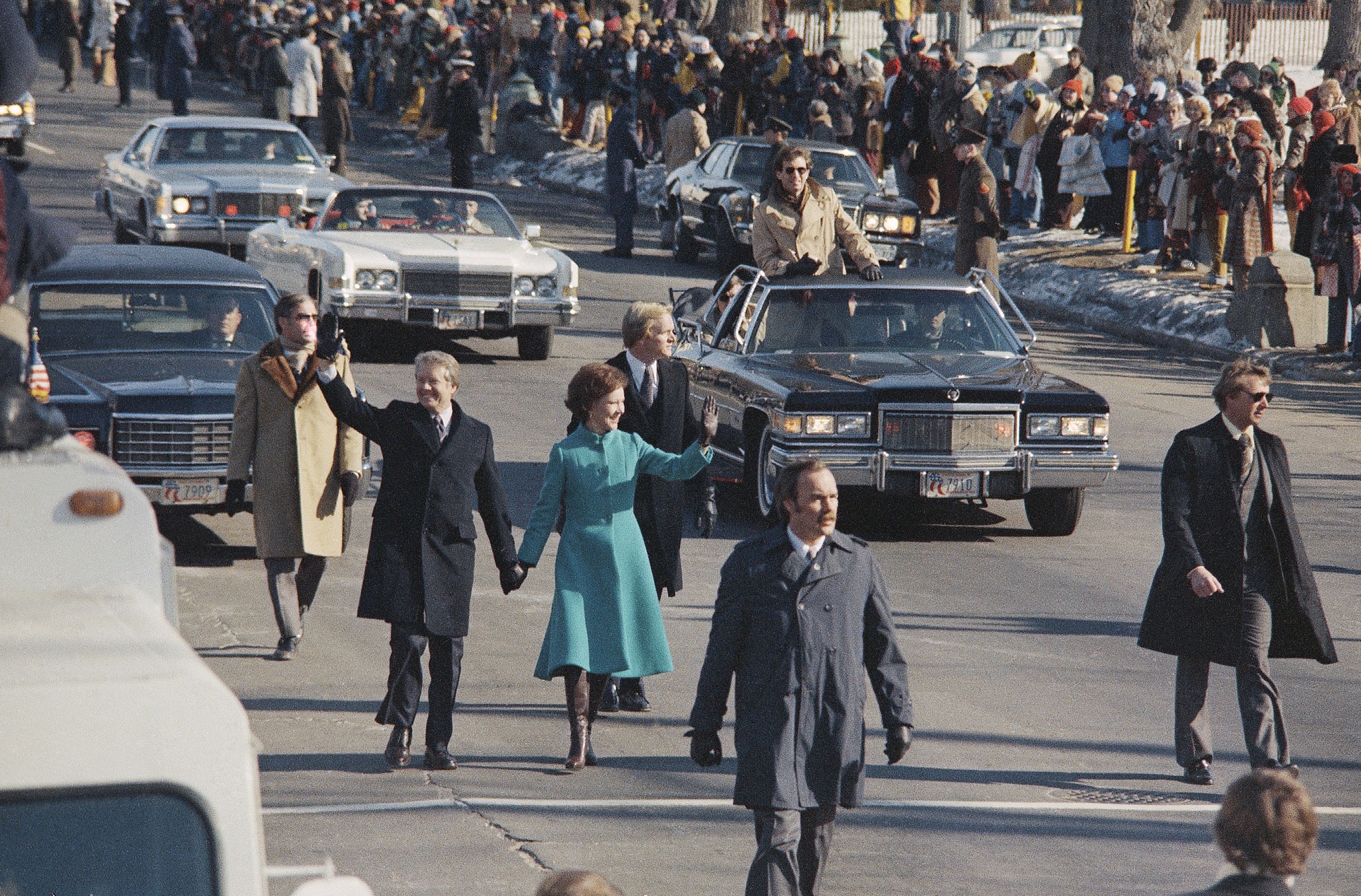 Jimmy Carter and Rosalynn Carter walking hand in hand in the street, waving to the crowd during their inaugural parade, surrounded by security and onlookers (© AP)