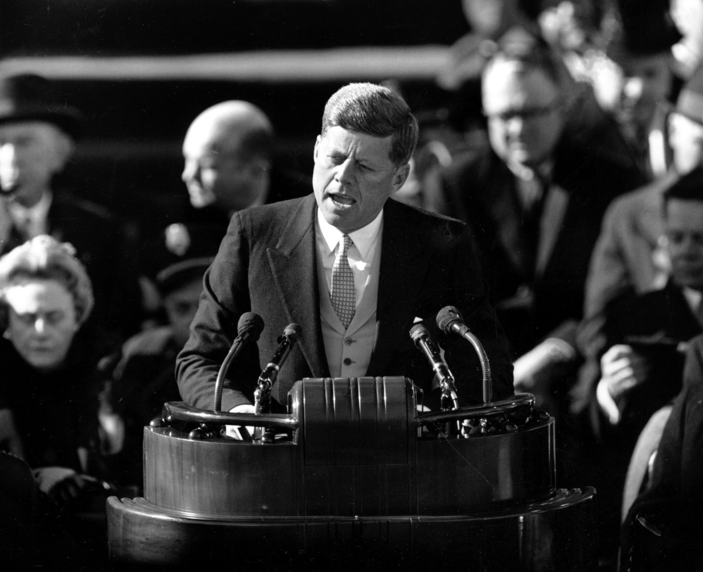 John F. Kennedy speaking  passionately into microphones, with dignitaries seated behind him (© AP)