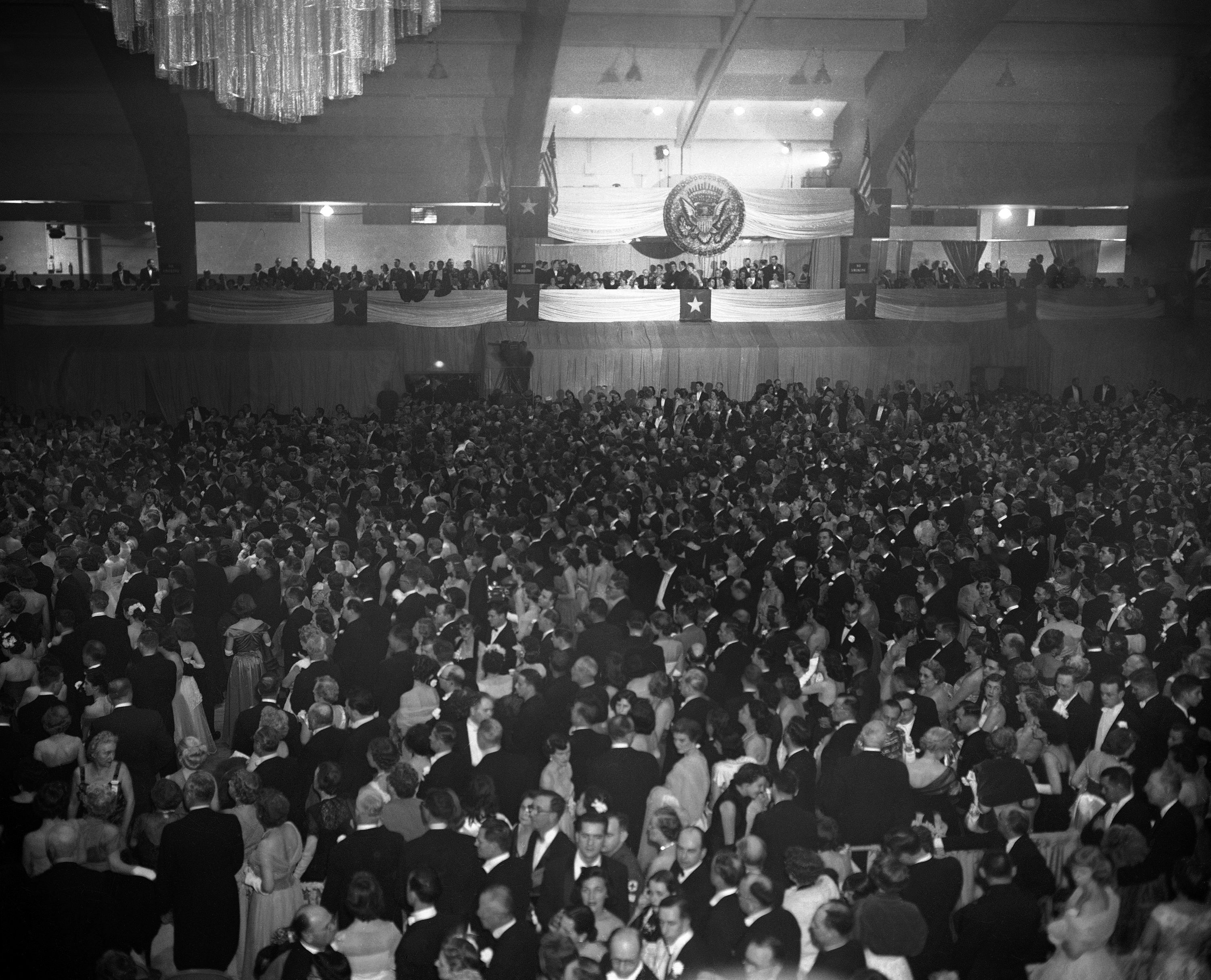 Black-and-white image of a packed ballroom during Dwight D. Eisenhower’s inaugural ball, with chandeliers and the presidential seal visible above (© AP)