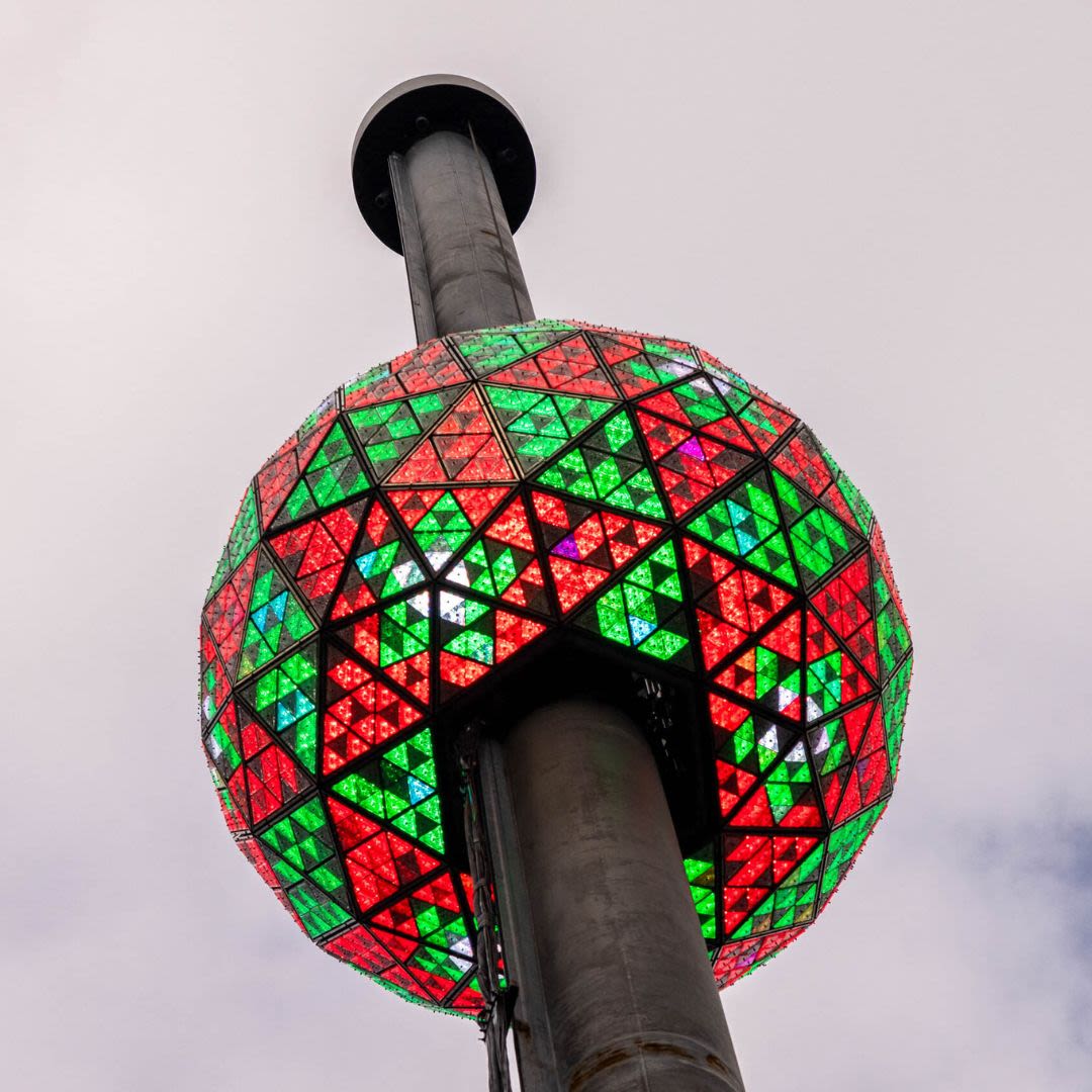 A close-up view of a New Year's Eve ball decorated with red and green illuminated triangular panels, set against a cloudy sky