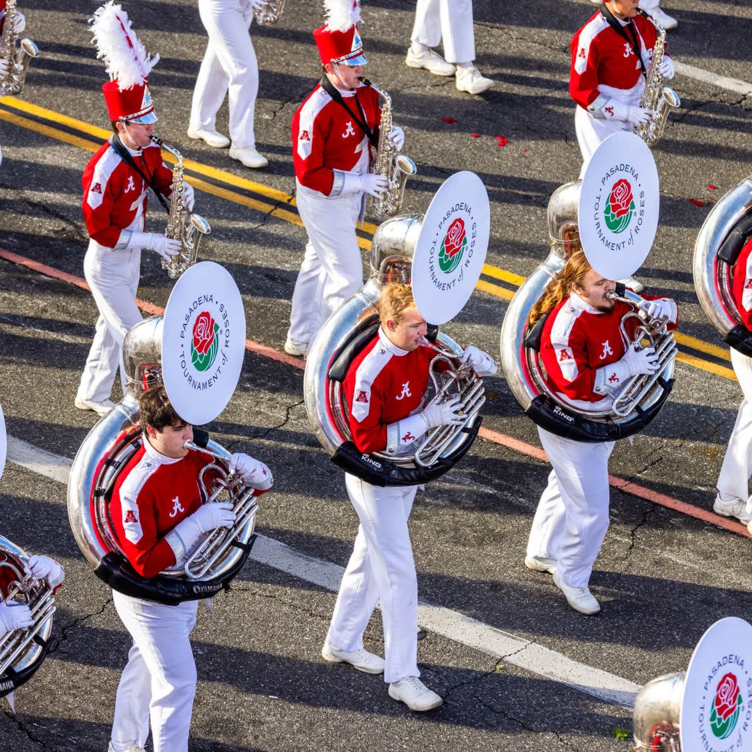 A marching band in a parade, wearing red and white uniforms, with sousaphones displaying “Pasadena Tournament of Roses”