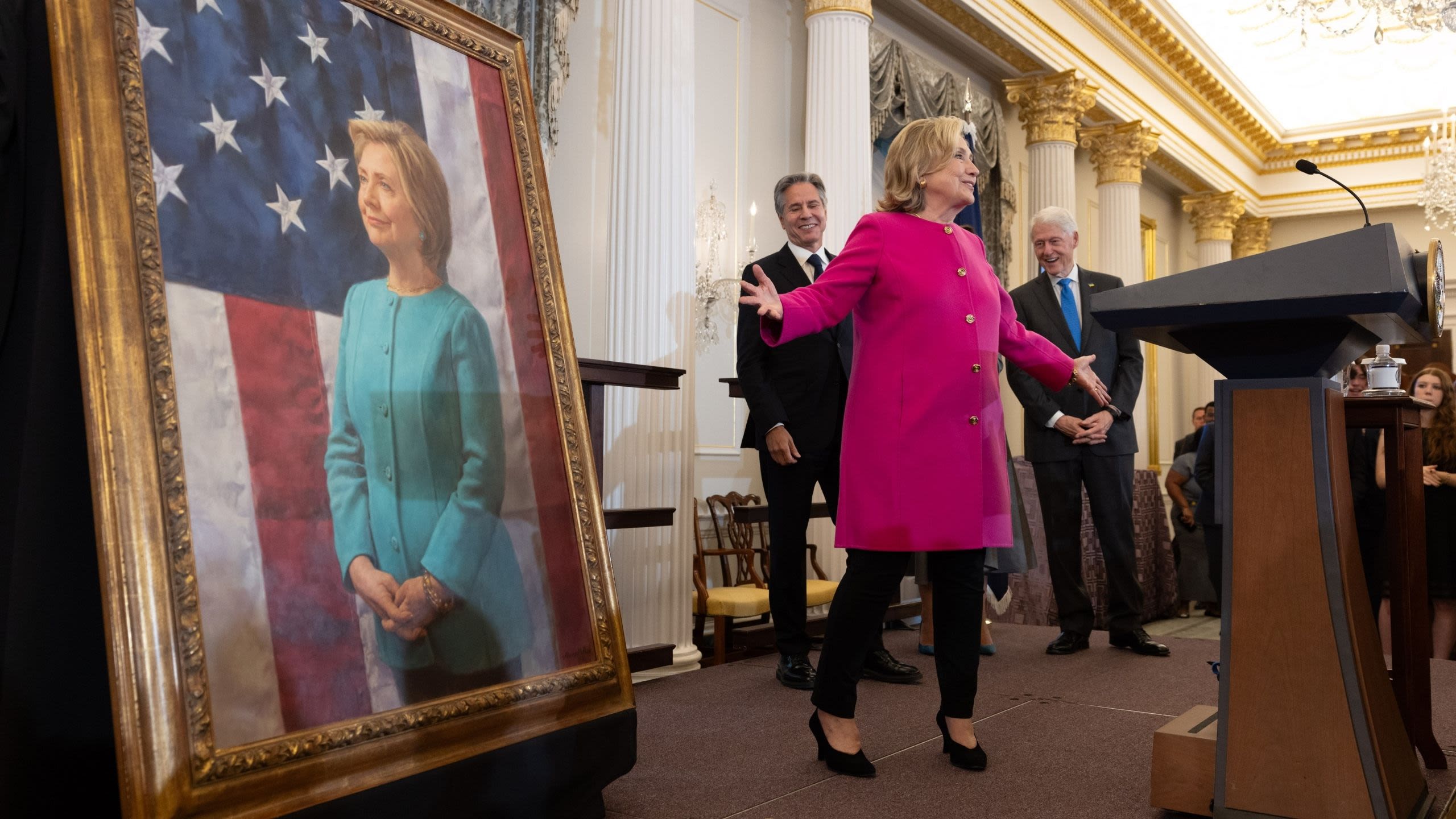 Hillary Clinton standing with arms outstretched in front of her official portrait