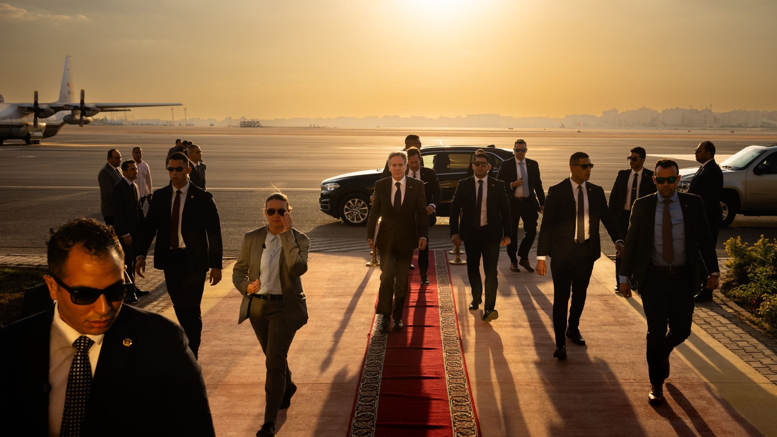 Secretary Blinken walking on a red carpet on a tarmac at sunset while surrounded by Diplomatic Security agents