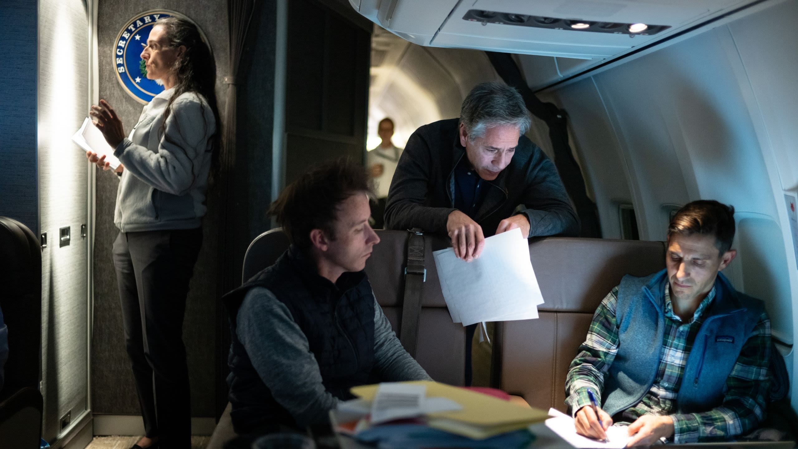 Secretary Blinken standing over two people sitting on a plane