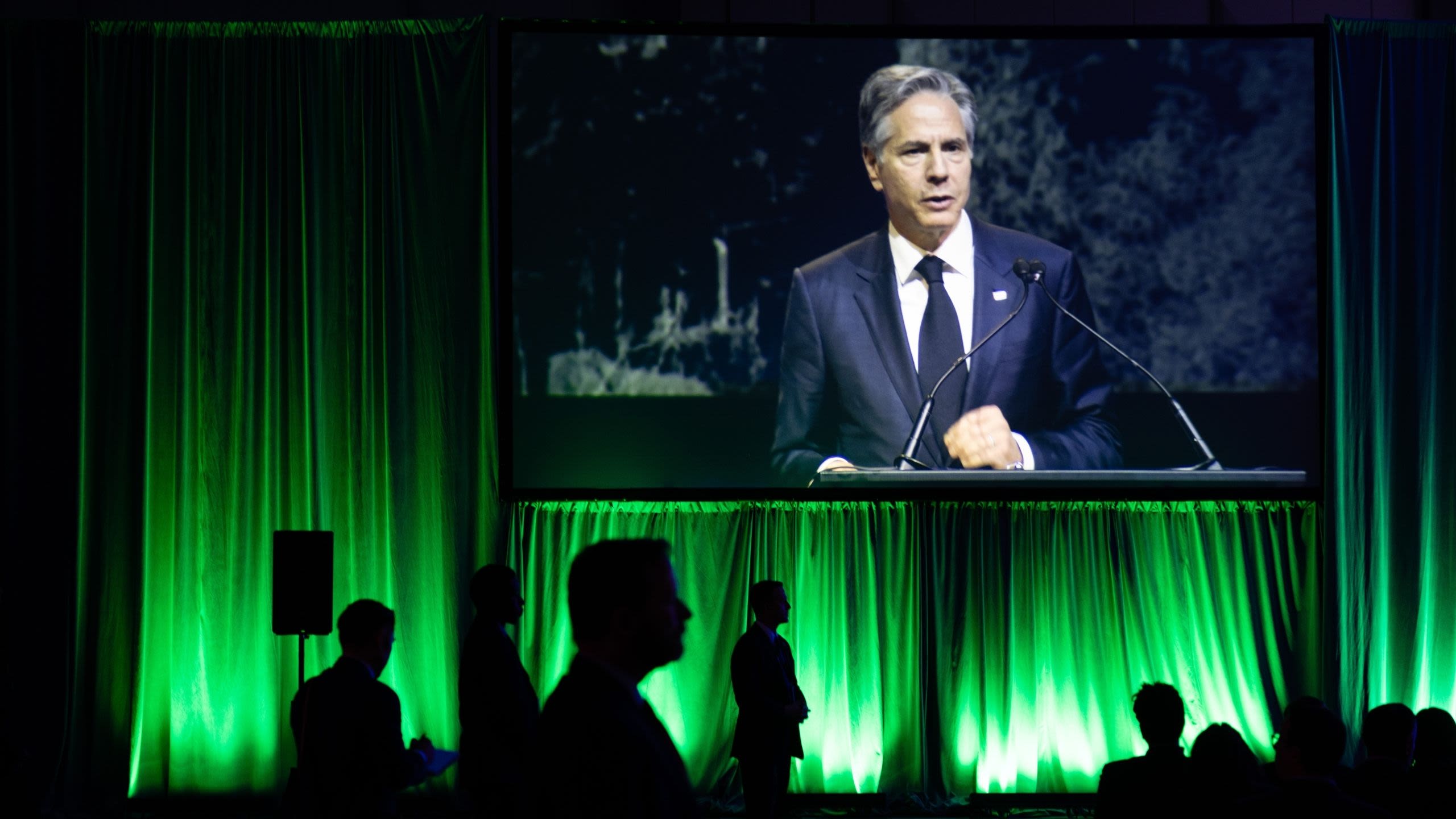 Projection screen showing Secretary Blinken speaking while security officers stand underneath