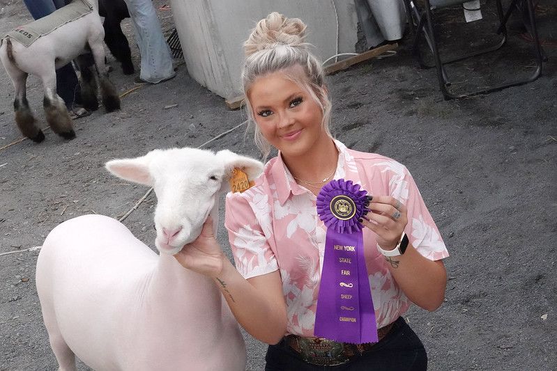 Young woman holding a champion ribbon next to a sheep (© Michael J. Okoniewski/New York State Fair)