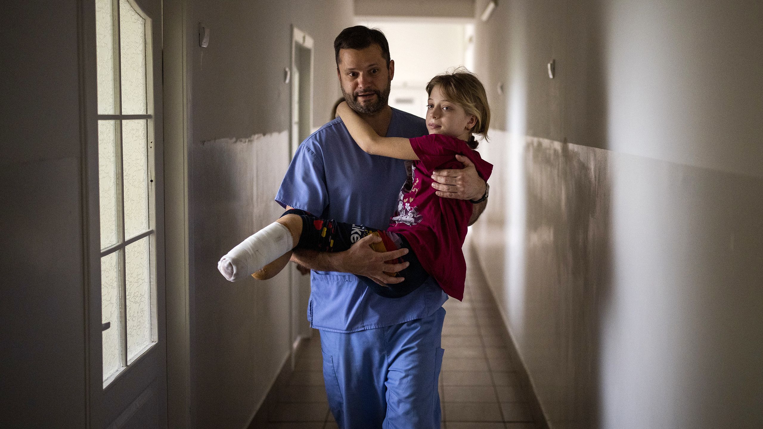Un homme dans un couloir, portant une petite fille amputée des pieds et ayant des bandages sur les jambes (© Emilio Morenatti/AP Photo)