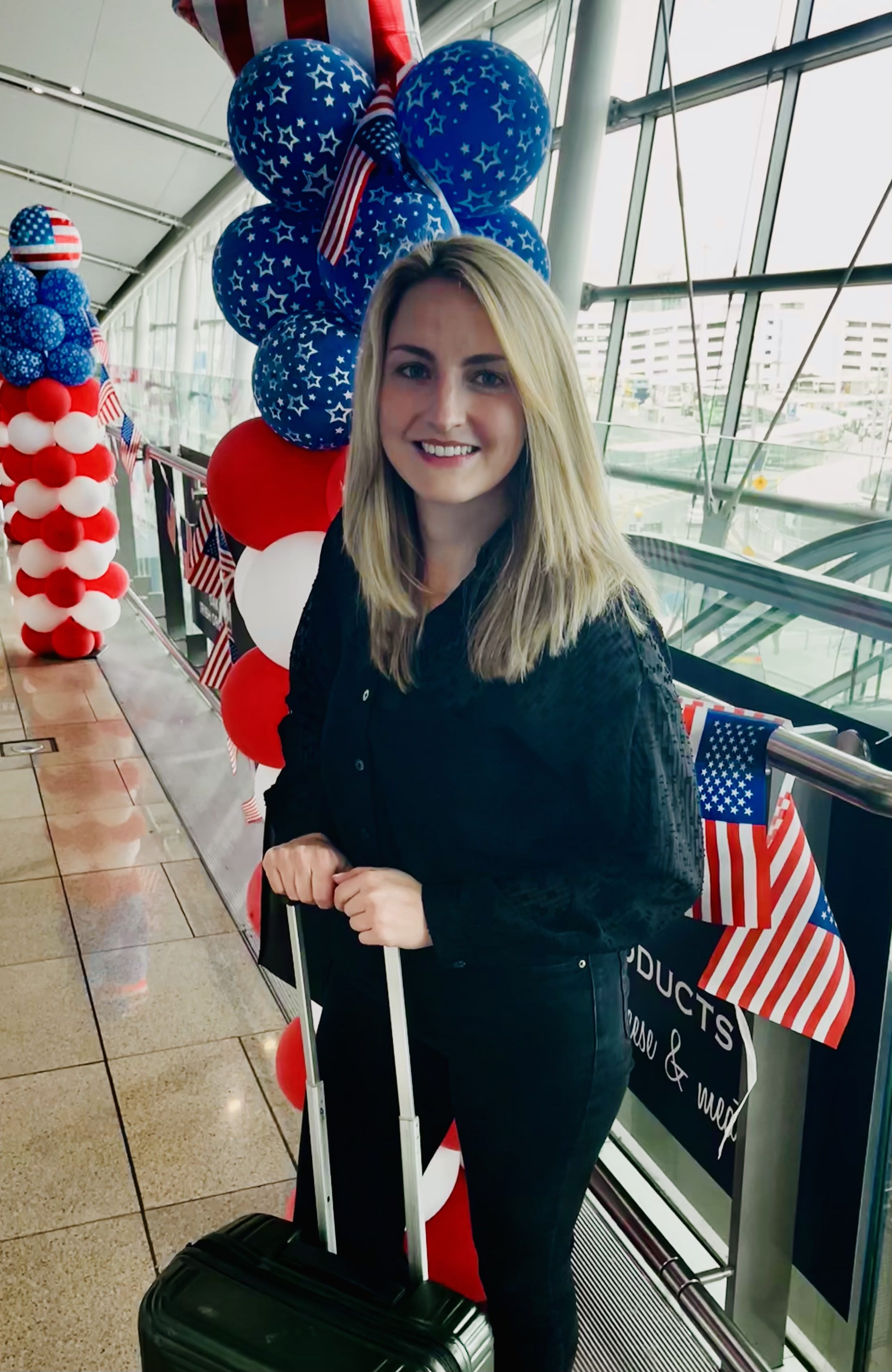 Woman standing with roller suitcase with red, white, and blue balloons behind her (Courtesy of Edith Delaney)