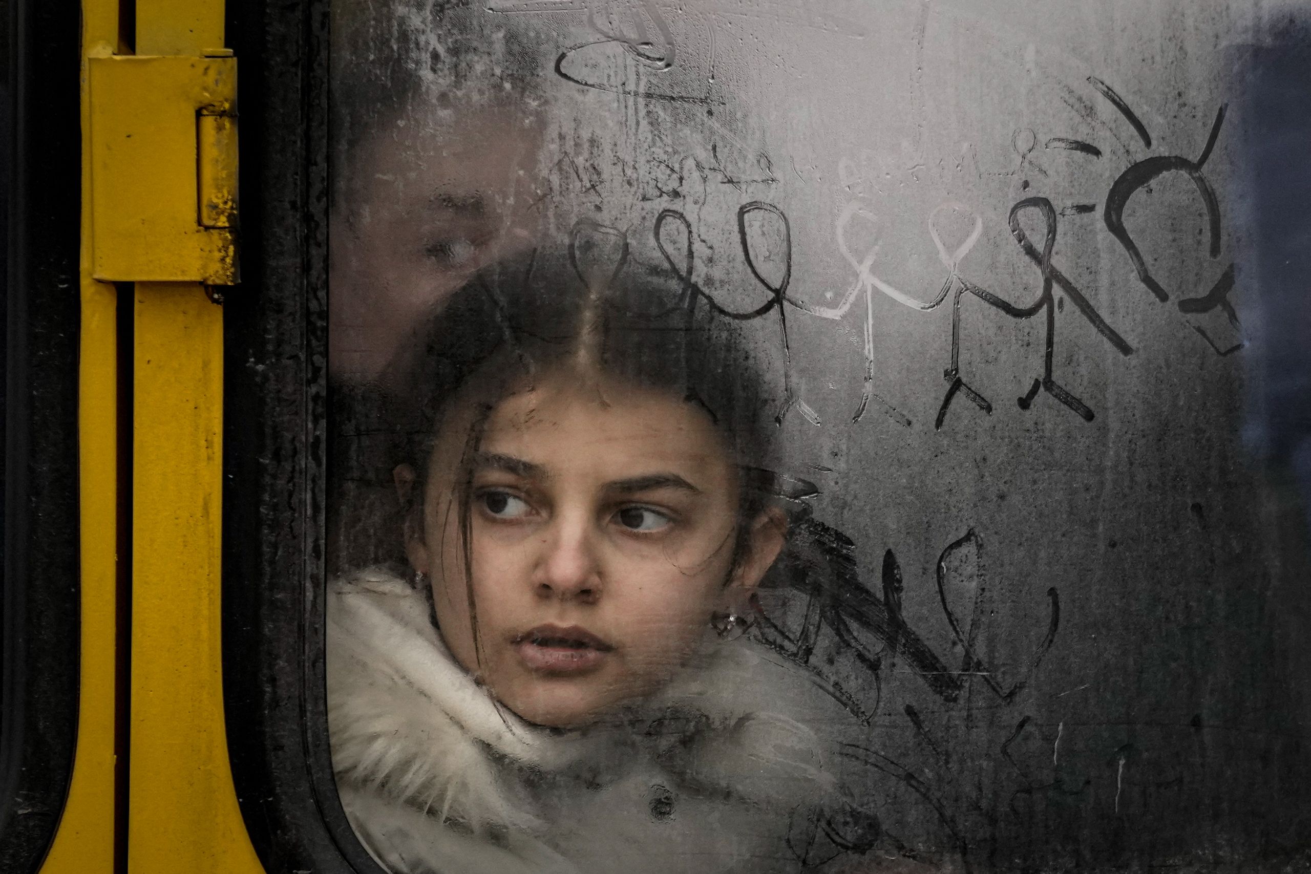 A child looks out a steamy bus window with drawings on it as civilians are evacuated from Irpin, on the outskirts of Kyiv, Ukraine, on March 9, 2022. (© Vadim Ghirda/AP Images)